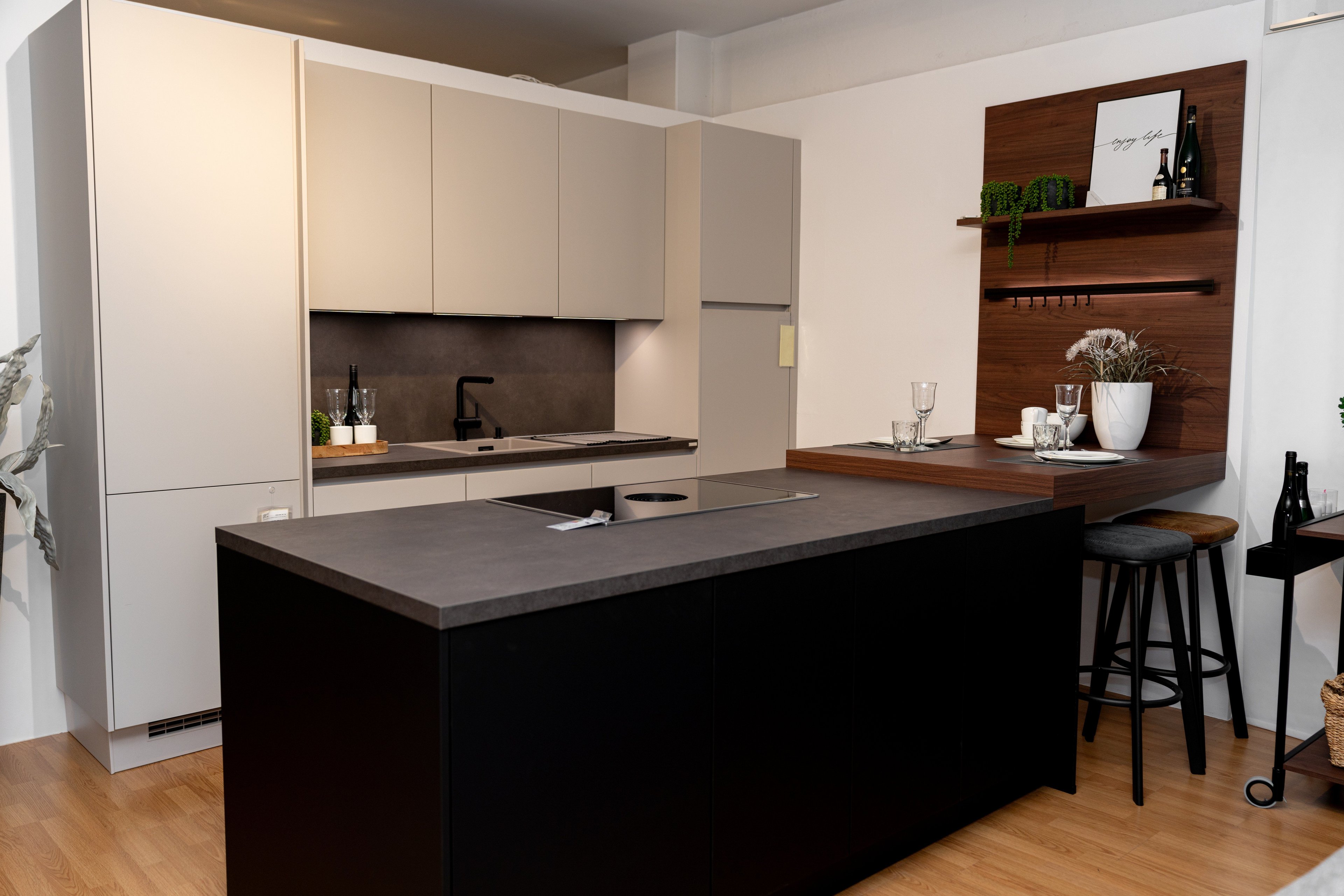 Moderne Küche mit beigen Oberschränken, schwarzer Kücheninsel mit grauer Arbeitsplatte und Nussbaum-Wandregal mit Pflanzen und Gläsern. Schwarze Barhocker stehen vor der Kücheninsel.