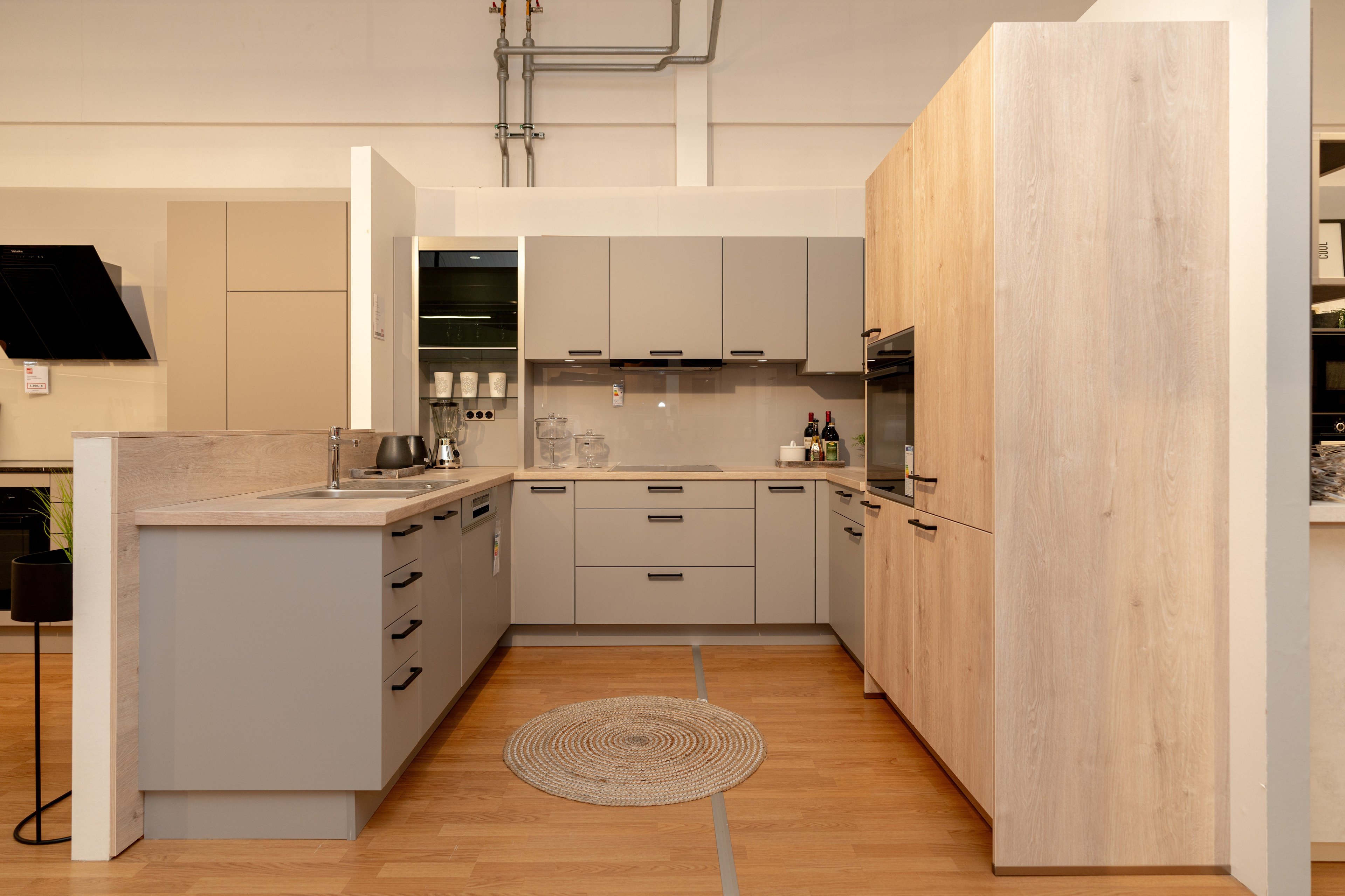 U-förmige Küche mit beigen Schränken und Holzoptik-Hochschrank rechts, Kücheninsel links mit Spüle. Runder geflochtener Teppich auf Parkett, industrielle Decke mit sichtbaren Leitungen.