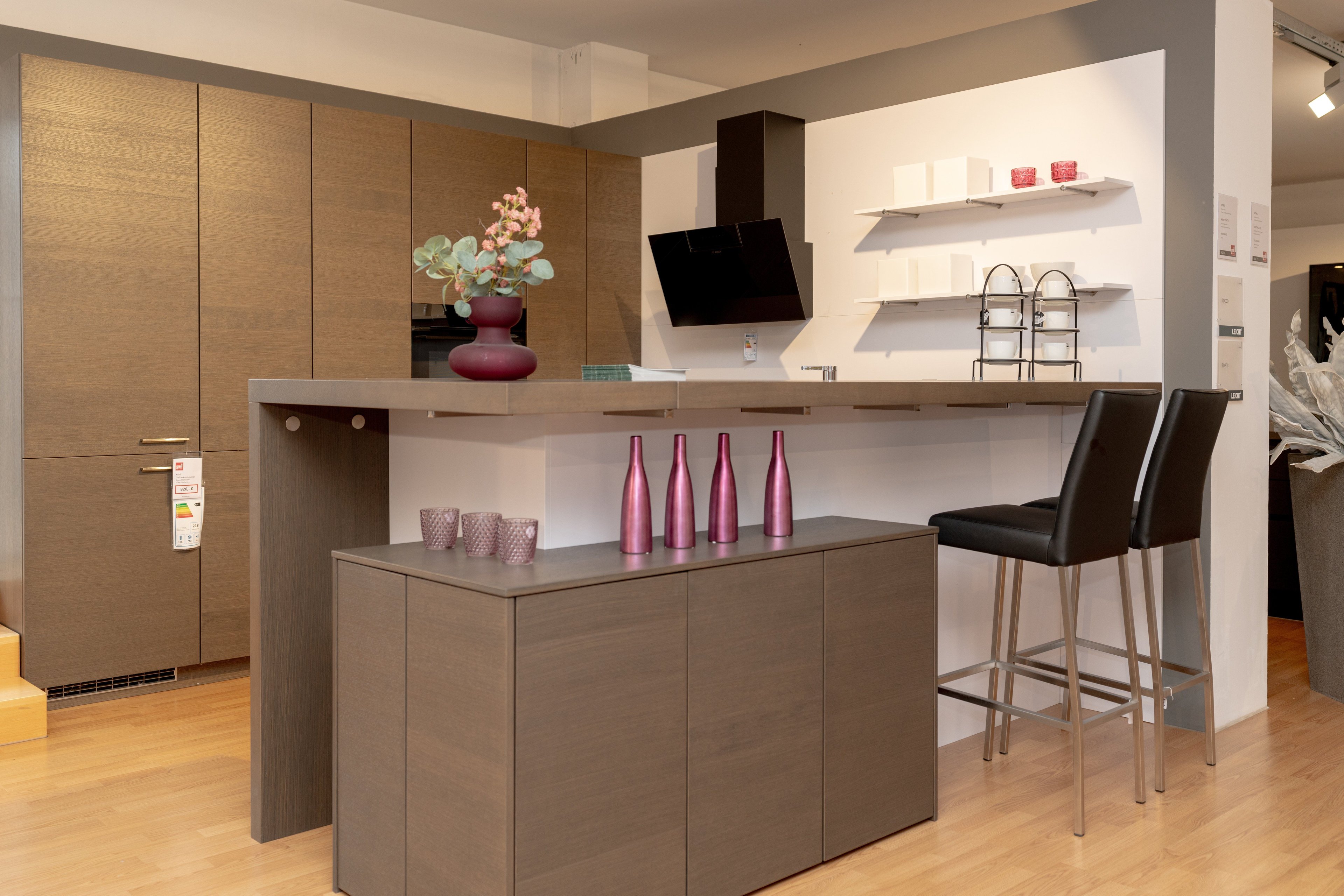 Moderne Küche mit holzfarbenen Oberschränken, grauer Kücheninsel mit rosa Dekorationsflaschen und schwarzen Barhockern. Weiße Wandregale und schwarze Dunstabzugshaube im Hintergrund.