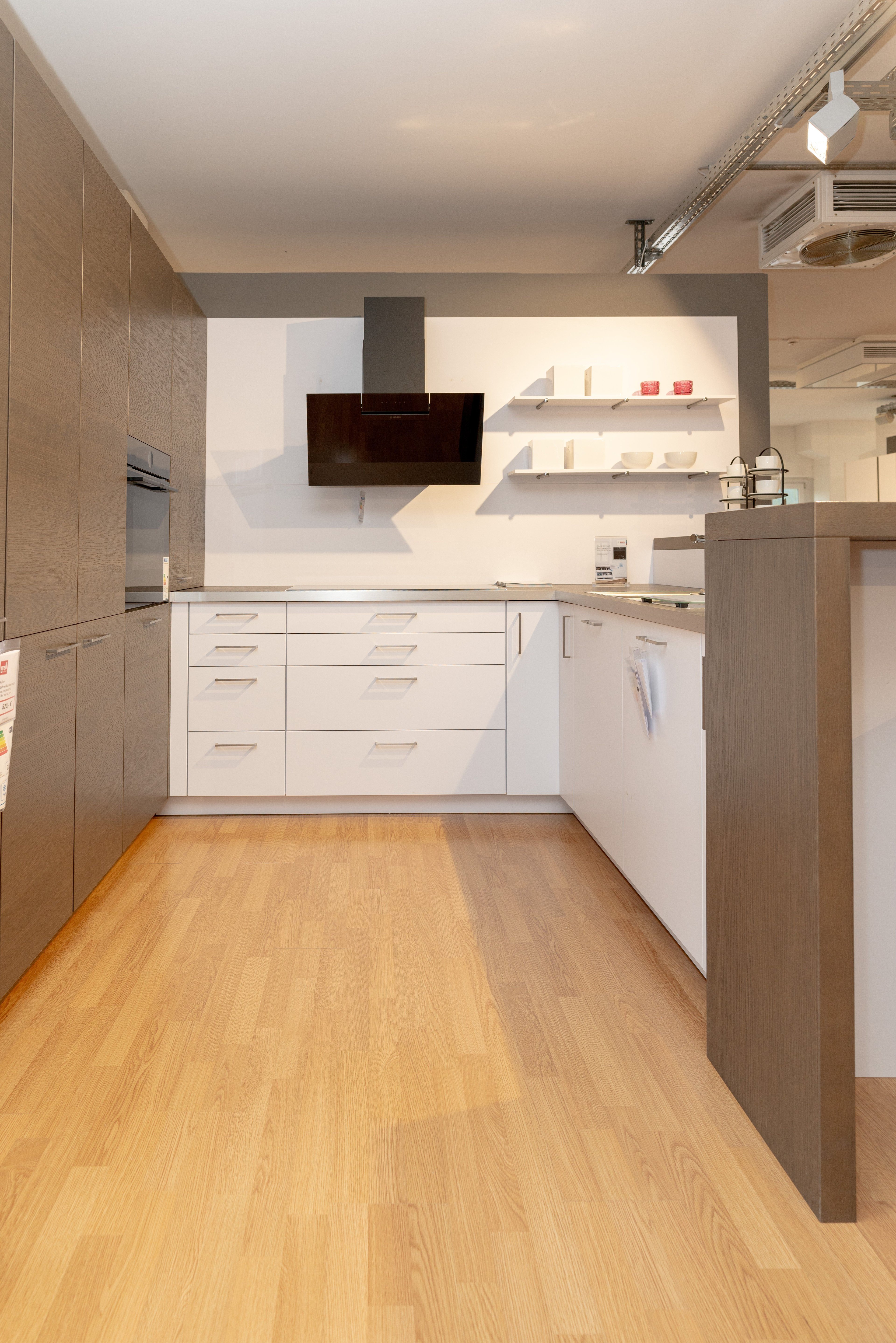 Moderne Küche mit holzfarbenen Oberschränken, grauer Kücheninsel mit rosa Dekorationsflaschen und schwarzen Barhockern. Weiße Wandregale und schwarze Dunstabzugshaube im Hintergrund.