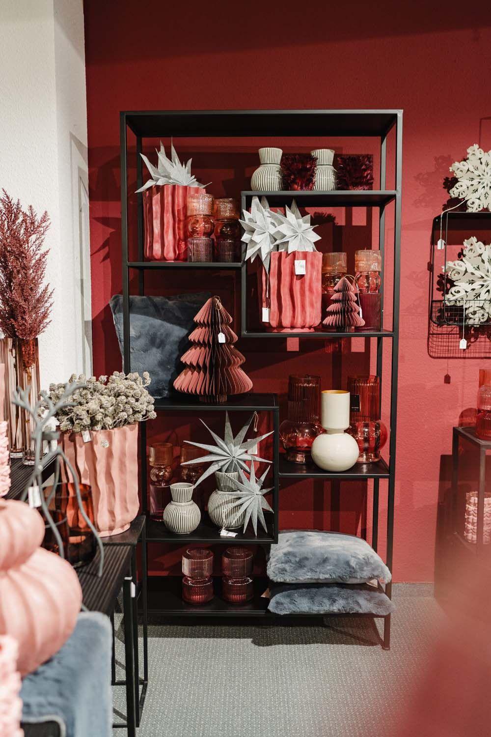 Regal mit roséfarbenen Vasen, Weihnachtsbäumen und weißen Sternen vor roter Wand.