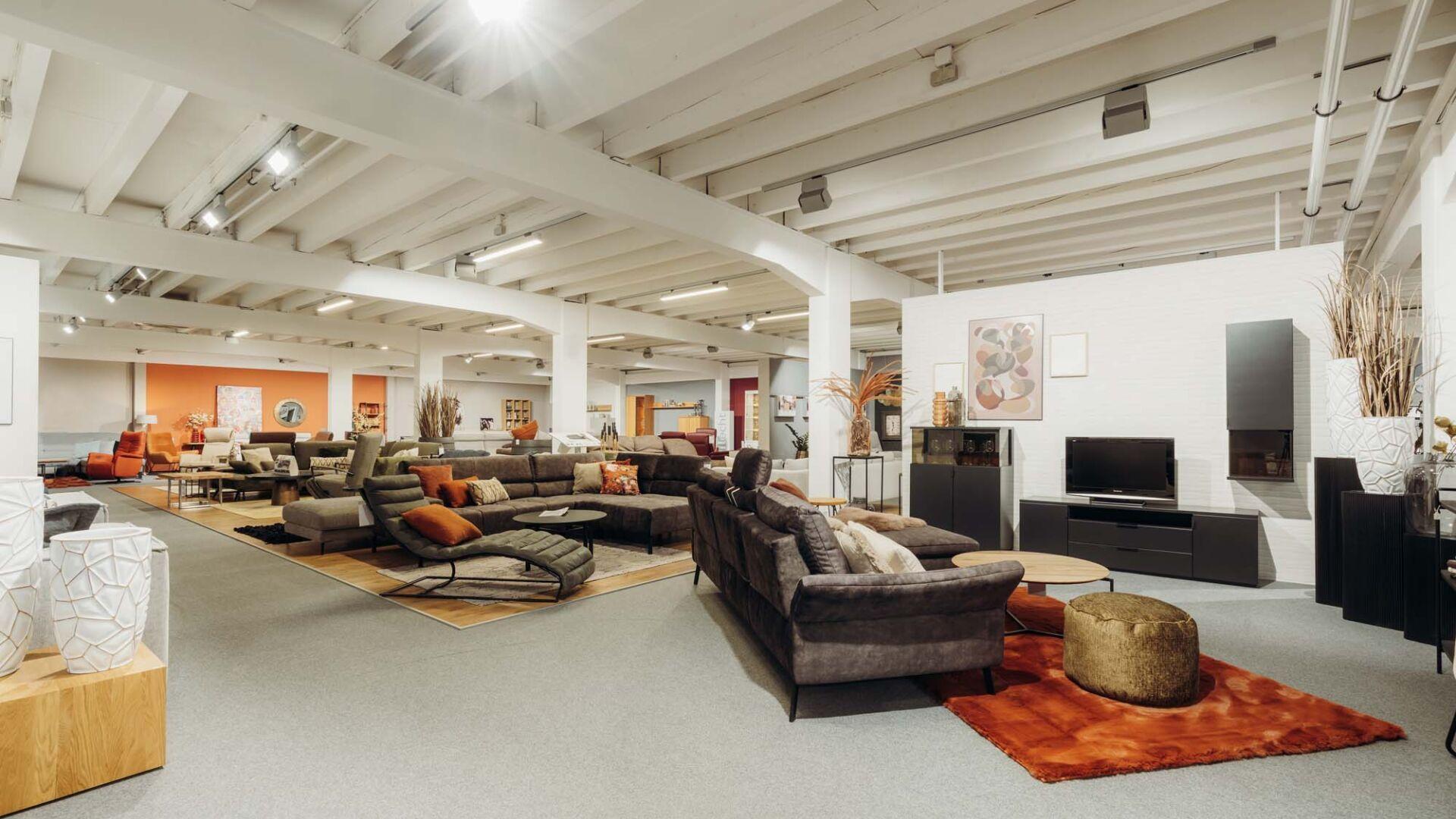 Großer Möbelshowroom mit offener Deckenstruktur, weißen Säulen und verschiedenen Wohnbereichen mit grauen und braunen Sofas, orangefarbenen Akzenten, TV-Möbeln und Teppichen in einem industriellen Loft-Ambiente. Großer Möbelshowroom mit offener Deckenstruktur, weißen Säulen und verschiedenen Wohnbereichen mit grauen und braunen Sofas, orangefarbenen Akzenten, TV-Möbeln und Teppichen in einem industriellen Loft-Ambiente.
