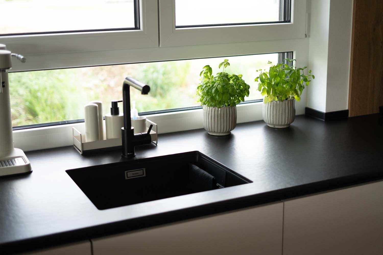 Schwarze Spüle mit Armatur am Fenster, Kräutertöpfen und dunkler Arbeitsplatte in moderner Küche