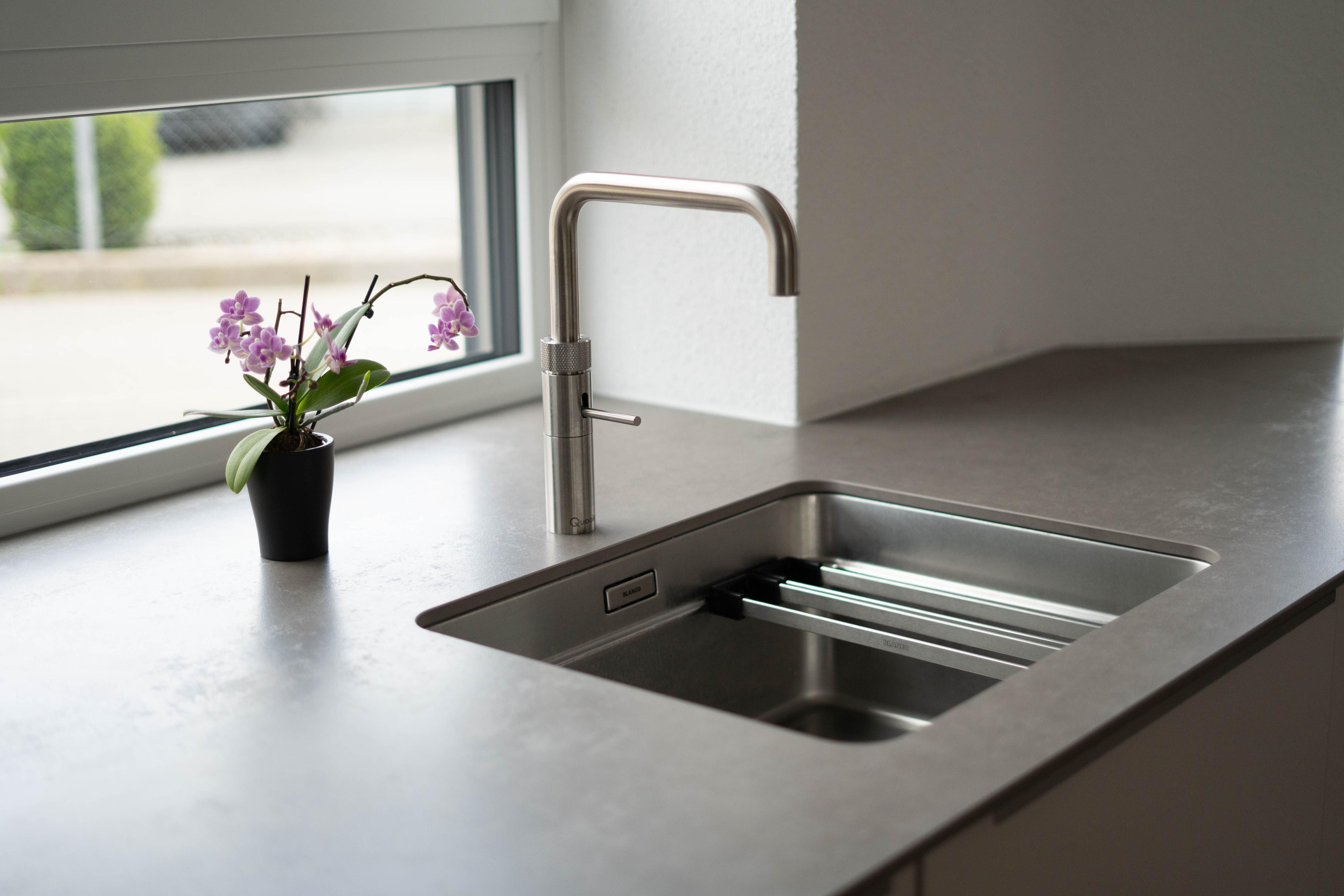 Spülenbereich am Fenster mit Edelstahlarmatur, grauer Arbeitsplatte und kleiner Orchidee als Dekoration