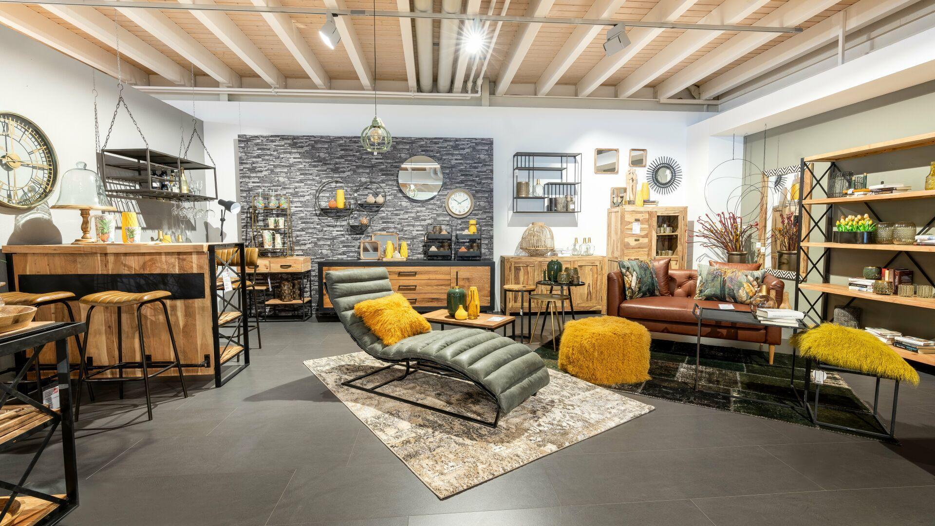 Moderner Möbel-Showroom im Industrial Style mit Holzbalkendecke, grauer Relaxliege, braunem Ledersofa und gelben Akzenten vor Steinwand.