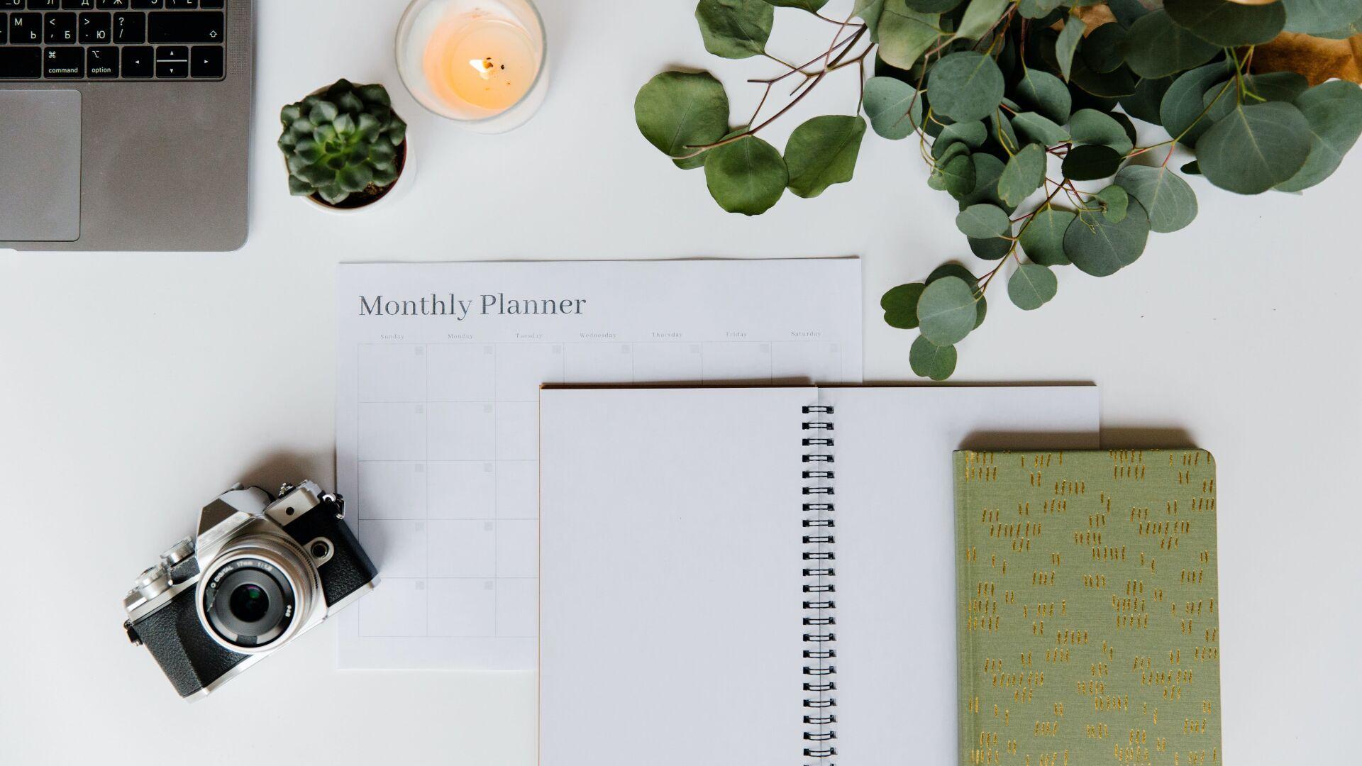 Schreibtisch von oben mit Laptop, Monatsplaner, aufgeschlagenem Notizbuch, Vintage-Kamera, brennender Kerze, kleiner Sukkulente und Eukalyptuszweigen auf weißem Untergrund. Schreibtisch von oben mit Laptop, Monatsplaner, aufgeschlagenem Notizbuch, Vintage-Kamera, brennender Kerze, kleiner Sukkulente und Eukalyptuszweigen auf weißem Untergrund.