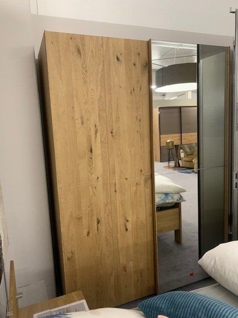 Moderner Kleiderschrank aus hellem Eichenholz mit Spiegeltür, teilweise geöffnet mit Blick ins Schlafzimmer.